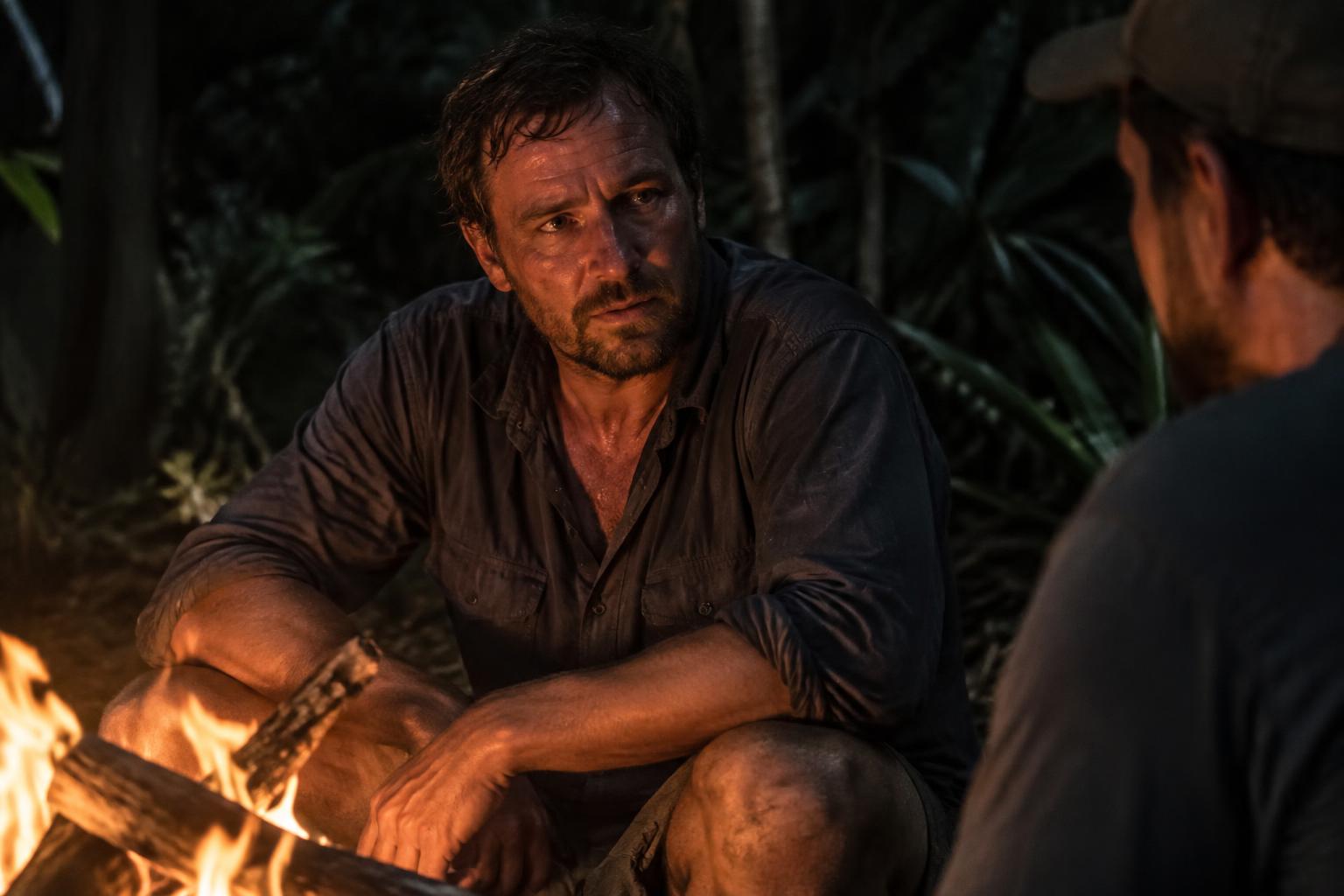 A man sitting by a campfire in conversation with another man at night